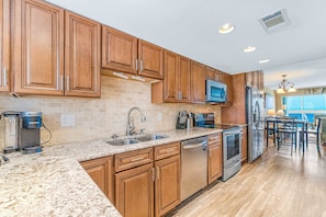 A kitchen with wooden cabinets, granite countertops, a dishwasher, a refrigerator, and a microwave. - Sunny, Gulf-front beach condo with central AC & a pool (Gulf Shores)