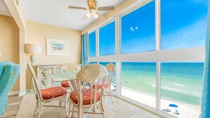 Una habitación con un ventanal grande con vista a una playa, sillas de mimbre, una mesa y un ventilador de techo.