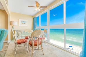 A room with a large window overlooking a beach, wicker chairs, a table, and a ceiling fan. - Sunny, Gulf-front beach condo with central AC & a pool (Gulf Shores)