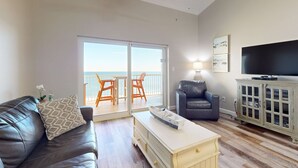 A living room with a sofa, a coffee table, a TV, a cabinet, and a balcony with a view. - Delightful beachfront condo w/ a furnished balcony, views, & a shared pool (Gulf Shores)