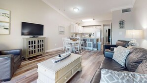 A living room with a TV, a dining area, and a kitchen in the background. - Delightful beachfront condo w/ a furnished balcony, views, & a shared pool (Gulf Shores)