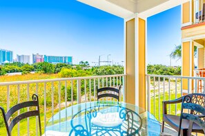 A balcony with a glass table and chairs, a white railing, and a view of a cityscape with buildings and greenery. - Snowbird-Friendly Condo w/Beach Access, Shared Outdoor Pool, Private Balcony (Orange Beach)
