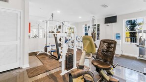 Ein Fitnessraum mit Trainingsgeräten, einem Fernseher und Blick auf Palmen.