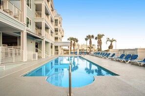A swimming pool with lounge chairs, a building with balconies, and palm trees. - Snowbird-Friendly Condo w/Beach Access, Shared Outdoor Pool, Private Balcony (Orange Beach)