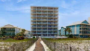 Un hotel de varios pisos con un paseo central que conduce a una zona de piscina, rodeado de vegetación junto a la playa y un muelle de madera.