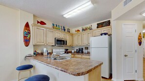 Una cocina con encimera de granito, armarios blancos, microondas, refrigerador y dos taburetes azules.