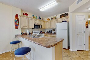 A kitchen with a granite countertop, white cabinets, a microwave, a refrigerator, and two blue bar stools. - Beachfront, corner condo w a furnished balcony, shared pool, & grilling area (Gulf Shores)