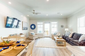 A spacious living room with a dining area, a gray sofa, a TV mounted on the wall, and a ceiling fan. - Luxurious, two-story cottage near the beach w a full kitchen & shared pool (Gulf Shores)