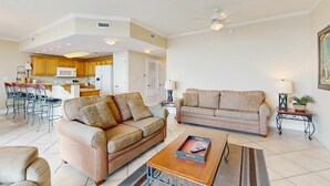 Ein Wohnzimmer mit zwei Sofas, einem Couchtisch und einem Essbereich mit Stühlen. Im Hintergrund ist eine Küche zu sehen.
