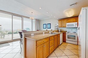 A modern kitchen with a large island, stainless steel appliances, wooden cabinets, and a view through large windows. - Gulf condo w/ private balcony, stunning views, pools, & sauna (Gulf Shores)