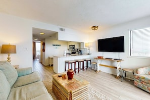 A living room with a sofa, a coffee table, a TV, and a kitchen area with a bar stool. - Condo w/ view of Little Lagoon, beach access, shared pools, hot tub, & pier (Gulf Shores)