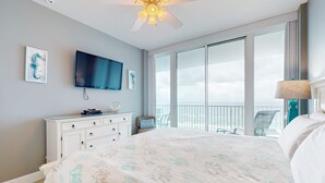 A bedroom with a bed, a TV, a dresser, a balcony with chairs, and a ceiling fan. - Beachfront condo with amazing views, multiple on-site pools & hot tubs (Gulf Shores)