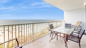 A balcony with a glass table and chairs, overlooking a beach with umbrellas and people. - Beachfront condo with amazing views, multiple on-site pools & hot tubs (Gulf Shores)