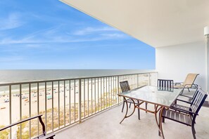 A balcony with a glass table and chairs, overlooking a beach with umbrellas and people. - Beachfront condo with amazing views, multiple on-site pools & hot tubs (Gulf Shores)