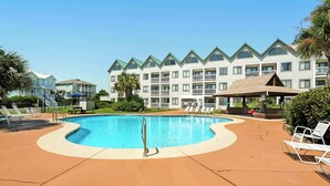 A swimming pool with lounge chairs, a covered seating area, and a multi-story building with balconies in the background. - Cozy waterfront getaway with pools, hot tub & tennis courts (Fort Morgan)