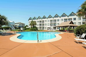 A swimming pool with lounge chairs, a covered seating area, and a multi-story building with balconies in the background.