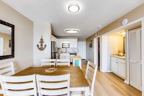A dining area with a wooden table and white chairs, a kitchen with white cabinets, and a hallway with a door. - Snowbird-friendly hotel suite with beach access, shared pool and dining (Fort Morgan)