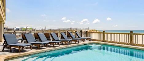 A pool with lounge chairs, a wooden fence, and a view of the beach.