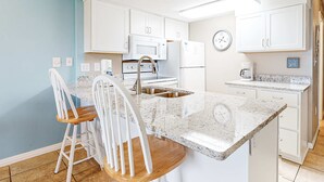 A kitchen with white cabinets, a microwave, a stove, a sink, and a coffee maker. There are two white bar stools with wooden seats. - Ground-floor condo with entertainment, shared pool, hot tub - near beach (Fort Morgan)