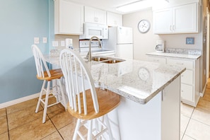 A kitchen with white cabinets, a microwave, a stove, a sink, and a coffee maker. There are two white bar stools with wooden seats. - Ground-floor condo with entertainment, shared pool, hot tub - near beach (Fort Morgan)