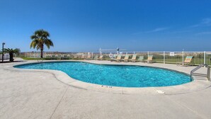 A swimming pool with a curved edge, a palm tree, and a white fence with lounge chairs. - Ground-floor condo with entertainment, shared pool, hot tub - near beach (Fort Morgan)