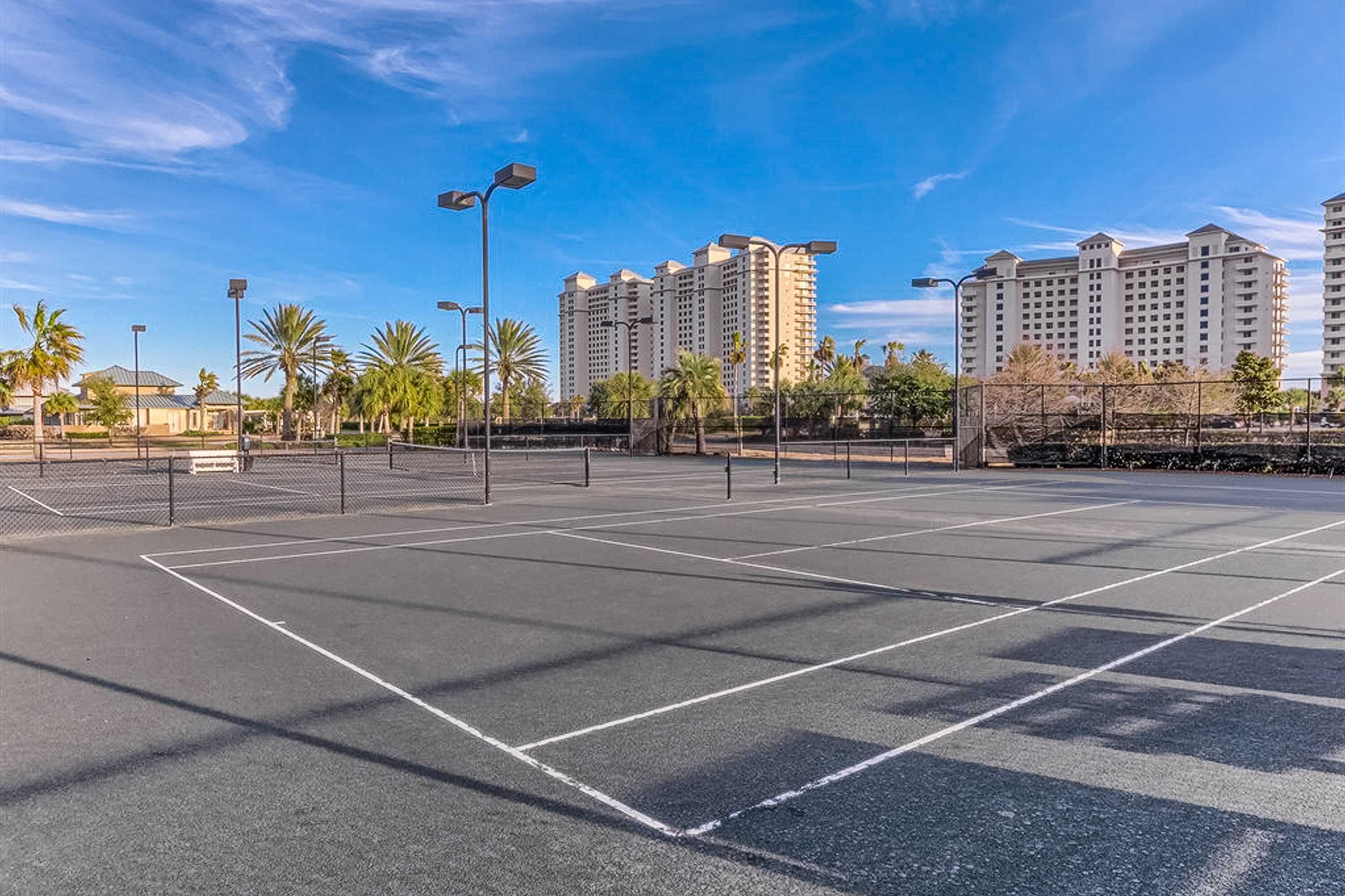 An outdoor tennis court with a clear sky, surrounded by palm trees and tall buildings.