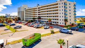 Un hotel de varios pisos con estacionamiento, un contenedor de basura verde y palmeras.