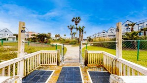 Una terraza de madera que conduce a una entrada con reja, con vista a edificios residenciales y palmeras.