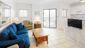 Ein Wohnzimmer mit blauem Sofa, einem Holztisch, einem Fernseher auf einem Standfuß, einer Küchenzeile inklusive Mikrowelle und einer Glastür zum Balkon.
