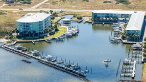 An aerial view of a marina with docked boats, a large building with a flat roof, and a smaller structure nearby. - Bay View Condo w/ Deck, Assigned Boat Slip, WiFi, AC & Shared Pools/Hot Tubs (Fort Morgan)