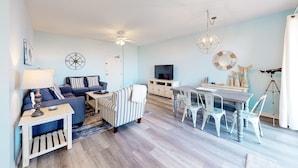 A living room with a sofa, a striped armchair, a dining table with chairs, a TV, and decorative items. - Bay View Condo w/ Deck, Assigned Boat Slip, WiFi, AC & Shared Pools/Hot Tubs (Fort Morgan)