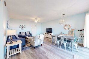 Ein Wohnzimmer mit einer Couch, einem gestreiften Sessel, einem Esstisch mit Stühlen, einem Fernseher und Dekorationselementen.