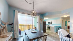 A dining area with a table and chairs, a kitchen in the background, and a balcony with a view. - Bay View Condo w/ Deck, Assigned Boat Slip, WiFi, AC & Shared Pools/Hot Tubs (Fort Morgan)