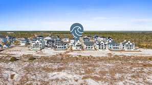 A coastal residential area with multiple houses, some with balconies and large windows, and a sandy beach in the foreground. - Beside Still Waters: Beachfront in MXB, Private Pool, Multiple Suites. (Mexico Beach)