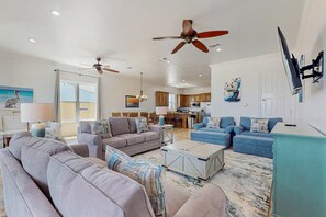 A spacious living room with sofas, a coffee table, ceiling fans, and a kitchen in the background. - Luxury Beachfront Duplex in Fort Morgan - Pool, Elevator and Wedding-Friendly (Fort Morgan)