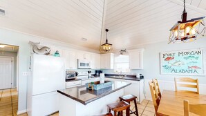 A kitchen with a central island, white cabinetry, a dining table with chairs, and a hanging light fixture. - Coastal Living Cottage on Mobile Bay with Private Beach Access (Fort Morgan)