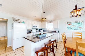 A kitchen with a central island, white cabinetry, a dining table with chairs, and a hanging light fixture. - Coastal Living Cottage on Mobile Bay with Private Beach Access (Fort Morgan)