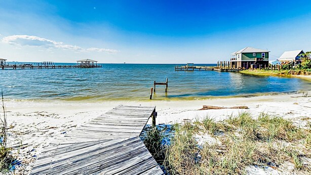 A beach with a wooden pier, a small structure, and a house with a green roof. - Coastal Living Cottage on Mobile Bay with Private Beach Access (Fort Morgan)