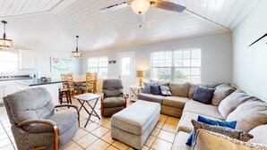 A spacious living room with a ceiling fan, a dining area with chairs, a sofa, and an ottoman. - Coastal Living Cottage on Mobile Bay with Private Beach Access (Fort Morgan)