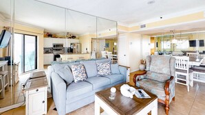 Ein Wohnzimmer mit einer Couch, einem Couchtisch und einem gemusterten Sessel. Im Hintergrund ist eine Küche erkennbar.