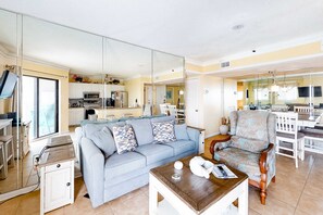 Ein Wohnzimmer mit einer Couch, einem Couchtisch und einem gemusterten Sessel. Im Hintergrund ist eine Küche erkennbar.