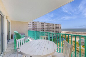 Un balcón con una mesa y sillas, vista a la playa y un edificio al fondo.