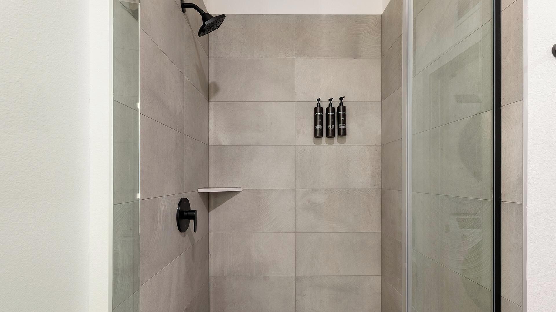 A shower with a glass door, a handheld showerhead, and two bottles of shampoo on a shelf.