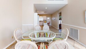 A dining area with a glass-top table surrounded by wicker chairs, a kitchen in the background, and a white railing on an upper level. - Dog-friendly house with private pool - dock and fishing pier available (Gulf Shores)