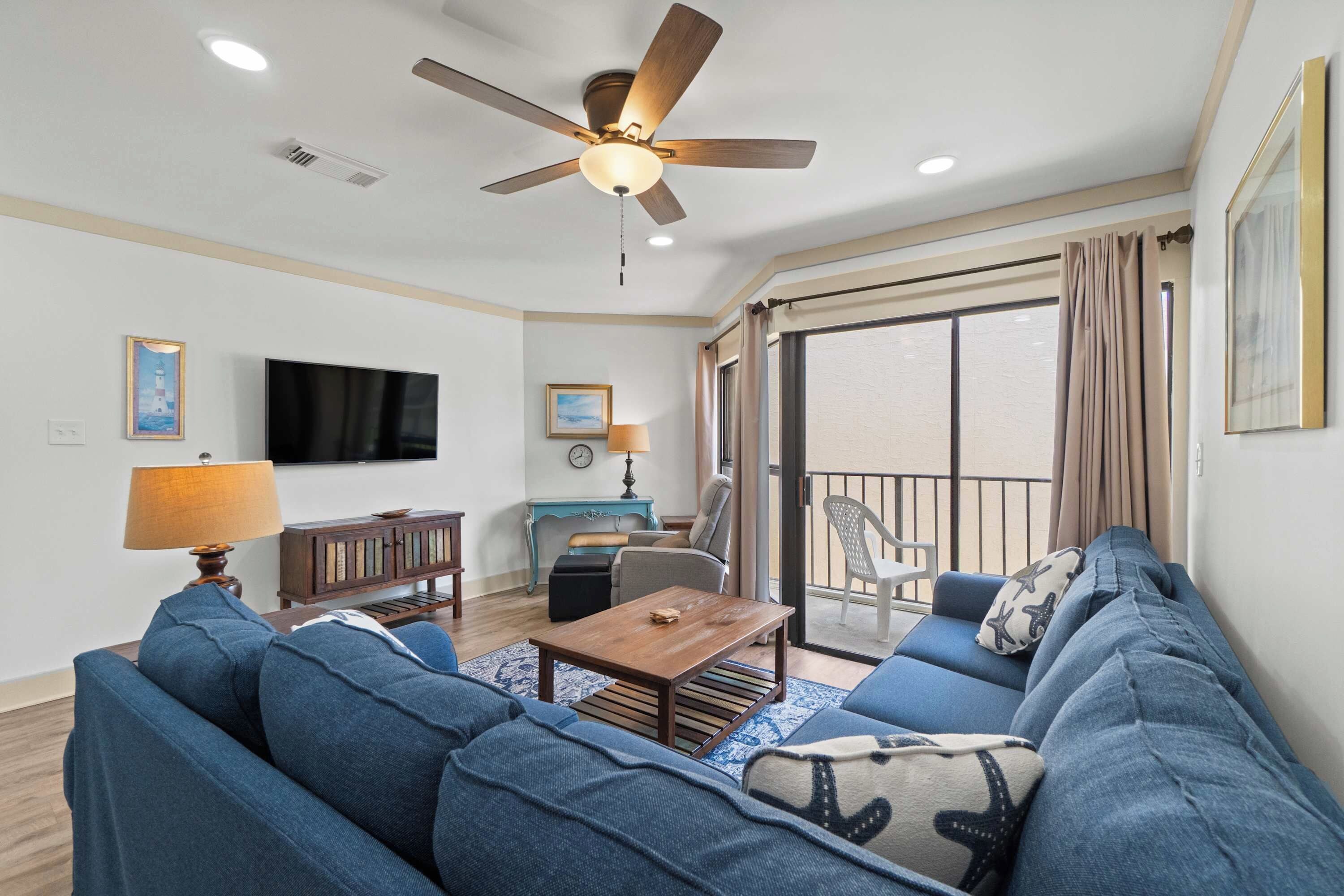 A living room with a ceiling fan, a flat-screen TV, a sofa set, a coffee table, and a view of the outside through a sliding glass door.