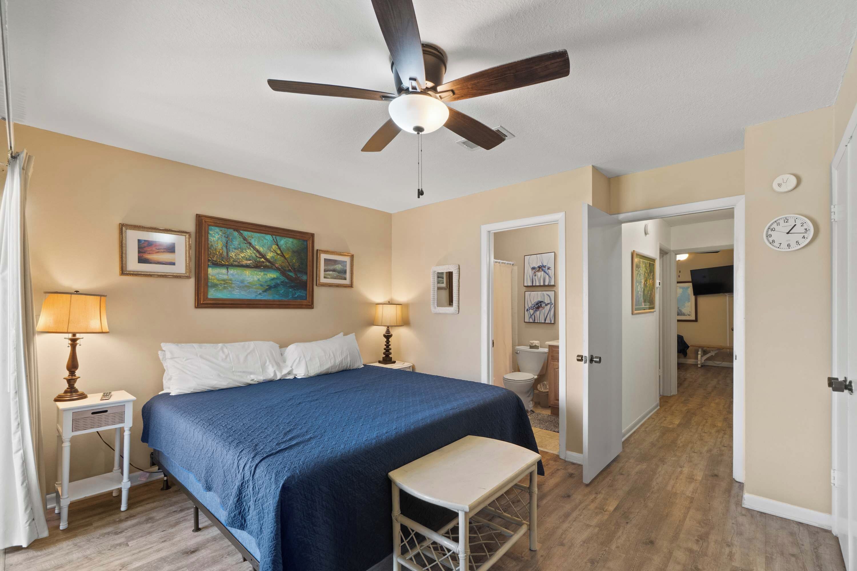 A bedroom with a bed, a nightstand, a ceiling fan, a window with curtains, and a door leading to another room.