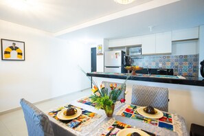 A dining area with a table set for two, a refrigerator, and a tiled backsplash in the background. - Park Del Prado #205 -ape em Cabedelo by Carpediem (Cabedelo)