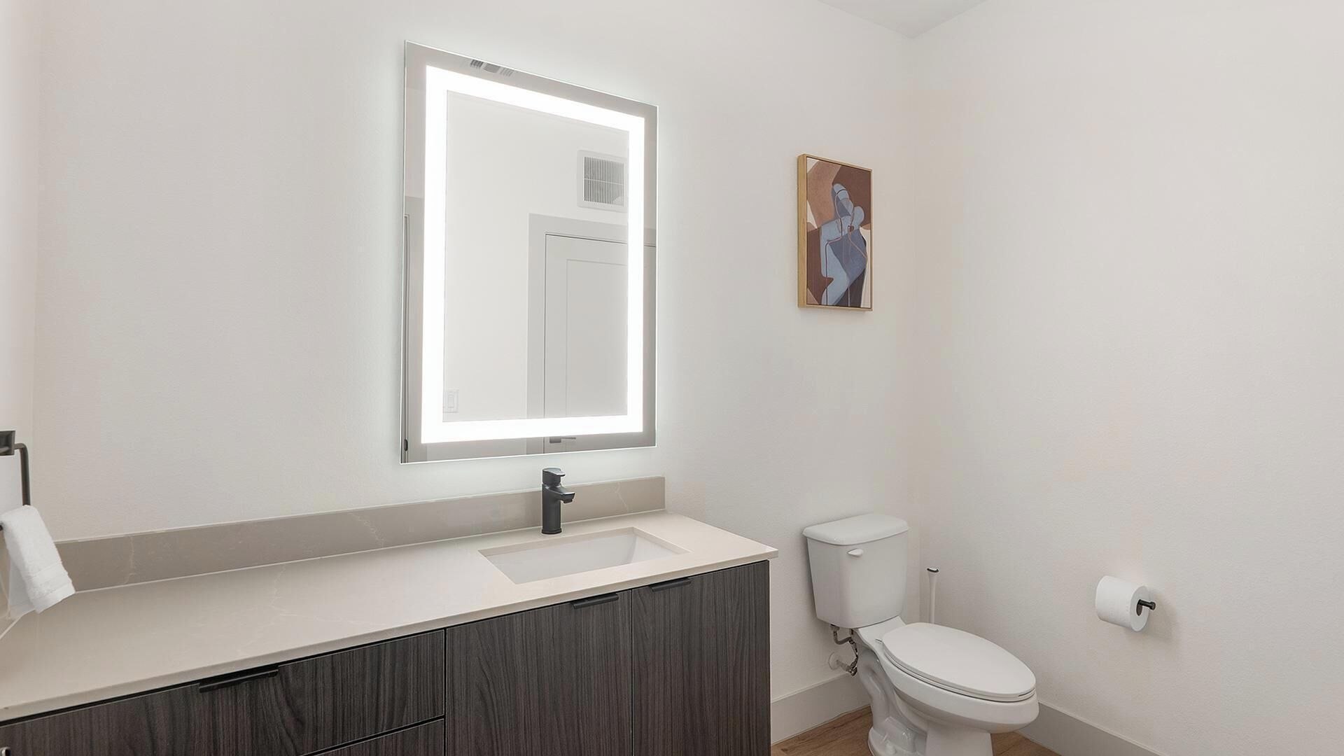 A bathroom with a mirror, sink, toilet, and a painting on the wall.
