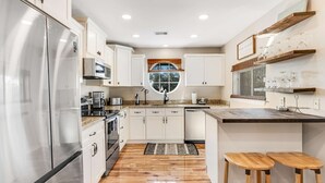 A modern kitchen with stainless steel appliances, white cabinetry, a central island with stools, and a large window. - Newly renovated, bright & chic Home with private deck, grill & near the beach (Fort Morgan)