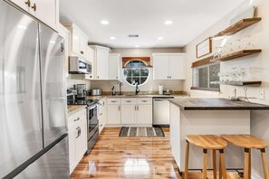 A modern kitchen with stainless steel appliances, white cabinetry, a central island with stools, and a large window. - Newly renovated, bright & chic Home with private deck, grill & near the beach (Fort Morgan)